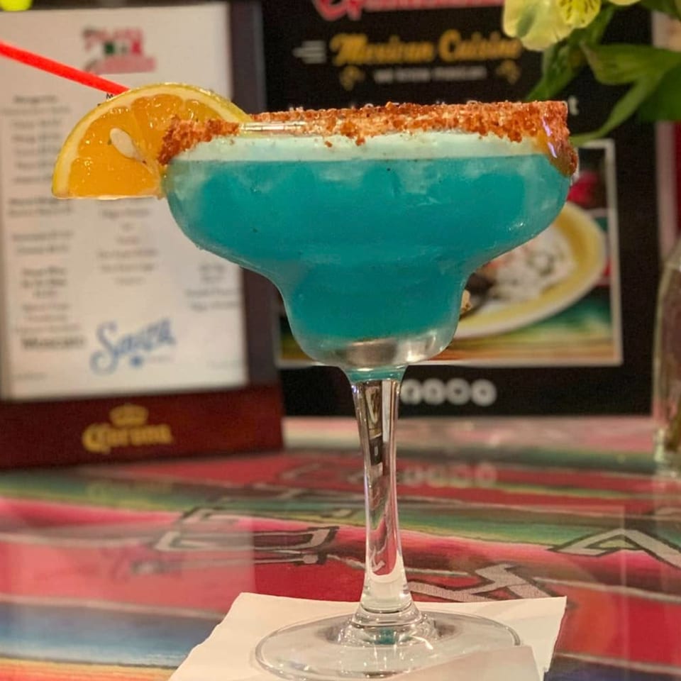 A vibrant blue margarita with a chili-lime rim and orange slice, on a colorful Mexican blanket.