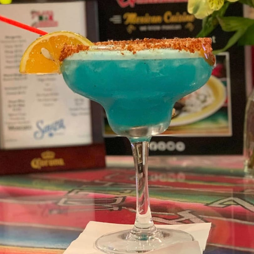 A vibrant blue margarita with a chili-lime rim and orange slice, on a colorful Mexican blanket.