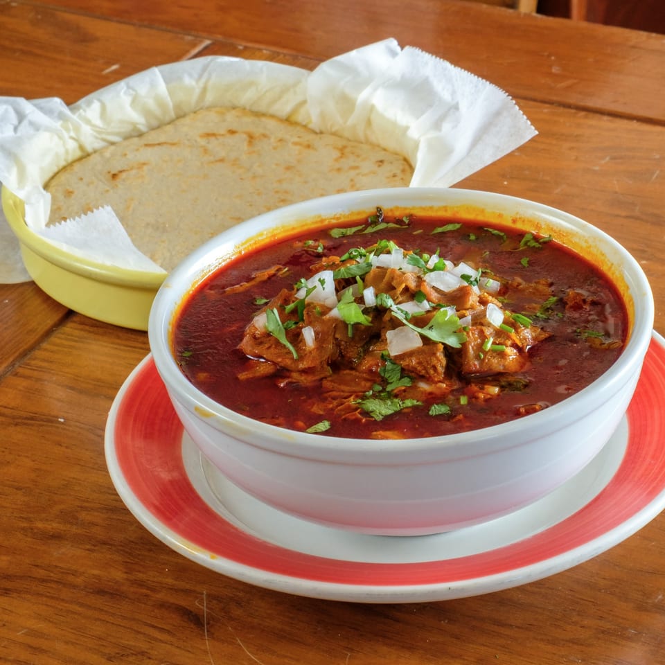 A bowl of red birria stew with cilantro and onions, served with tortillas.