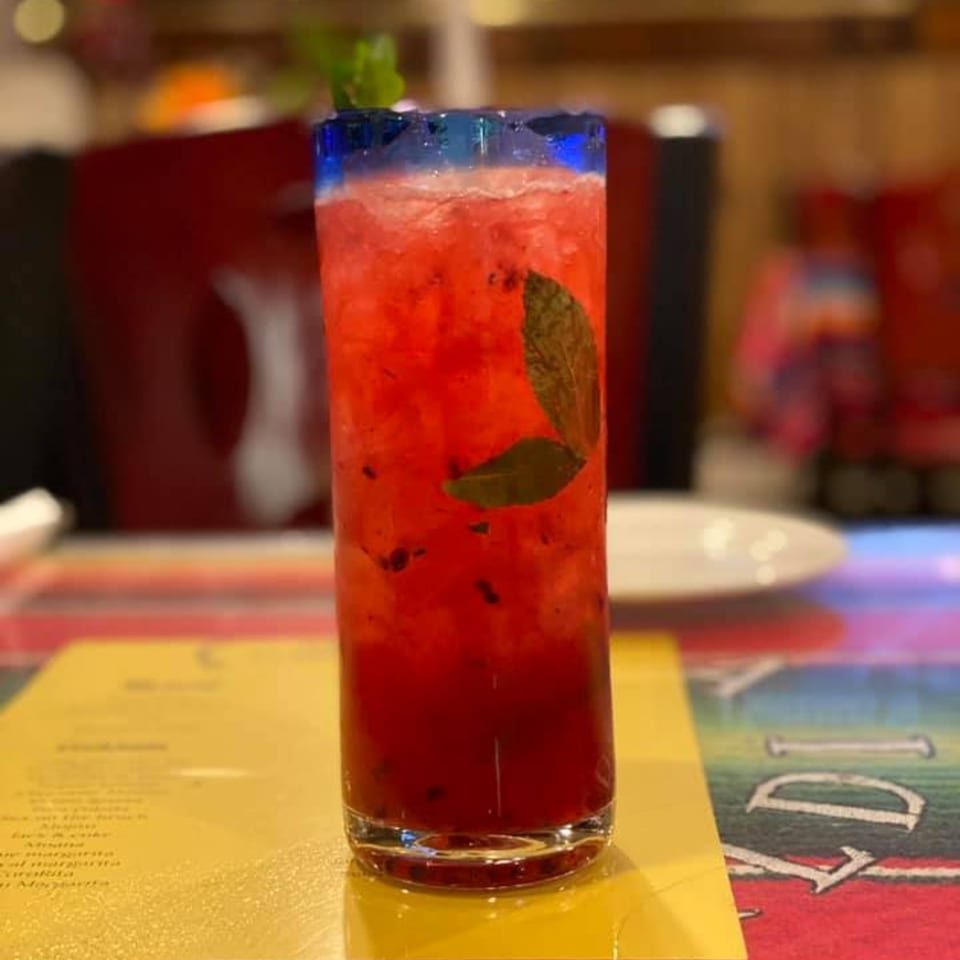A vibrant red mezcal cocktail with mint leaves and a blue rim sits on a colorful table.