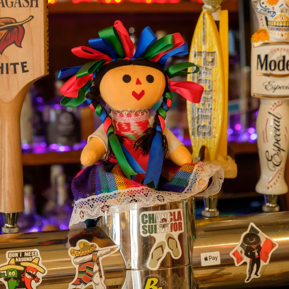 A colorful Mexican rag doll sits on a bar tap, surrounded by beer taps and stickers.