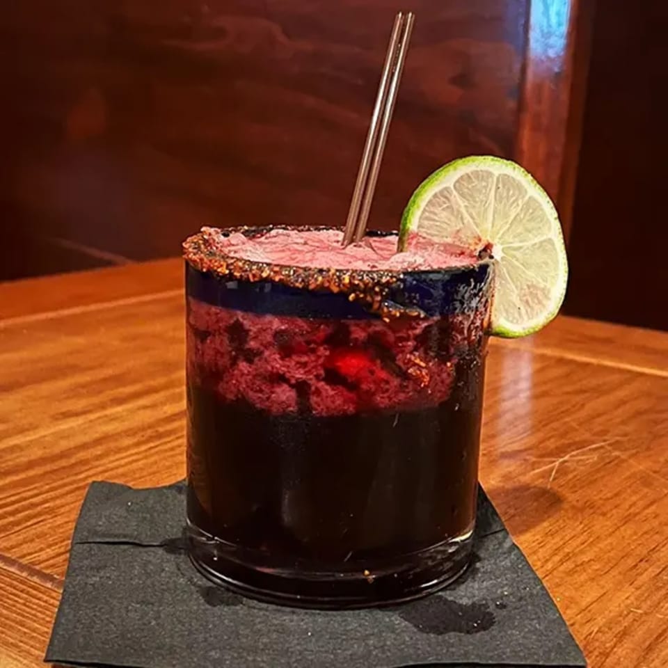 A dark red mezcal cocktail with a foamy top, chili-lime rim, and lime wedge on a wooden table.