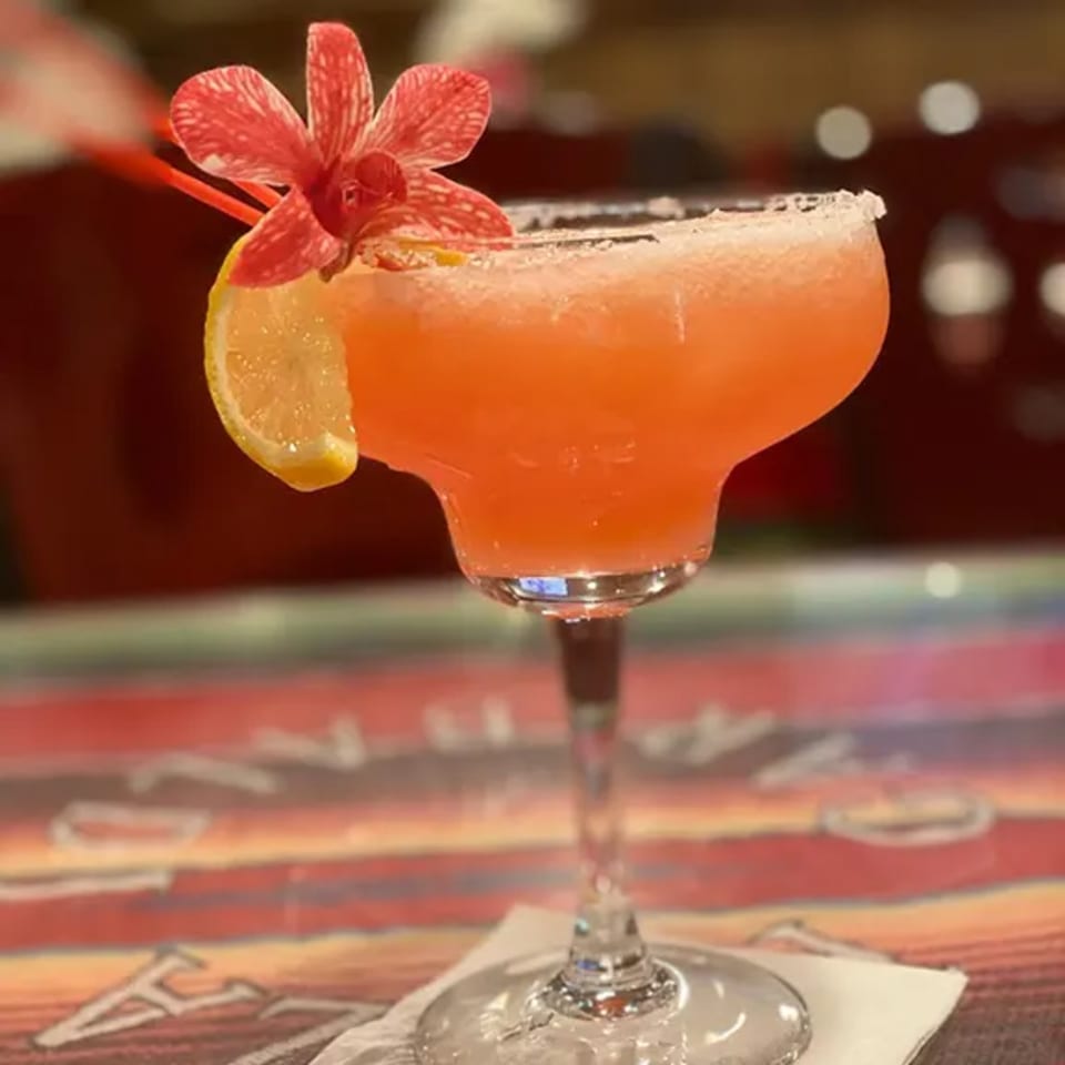 A vibrant orange margarita with a salted rim, garnished with a lemon slice and a red orchid.