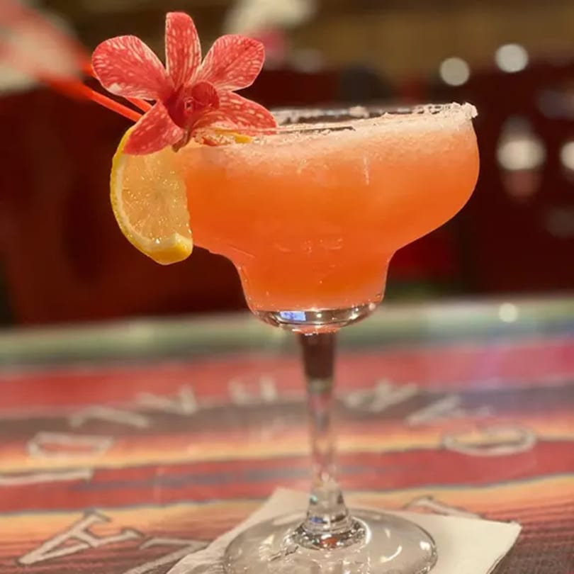 A vibrant orange margarita with a salted rim, garnished with a lemon slice and a red orchid.