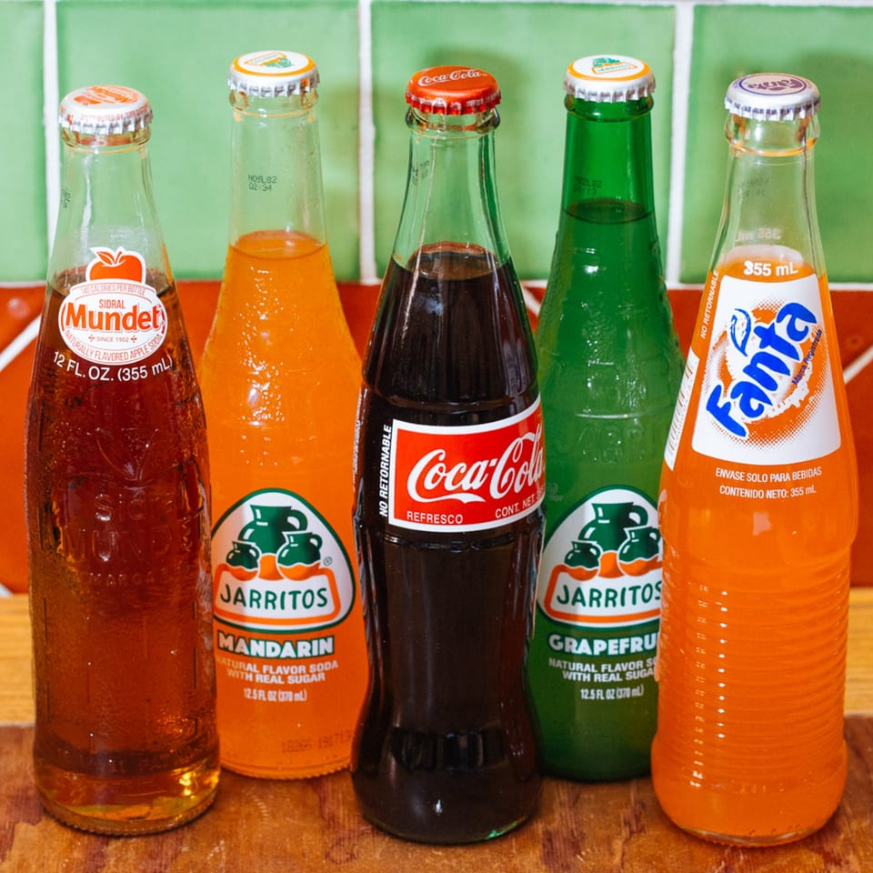 A row of five Mexican sodas: Sidral Mundet, Jarritos Mandarin, Coca-Cola, Jarritos Grapefruit, and Fanta.