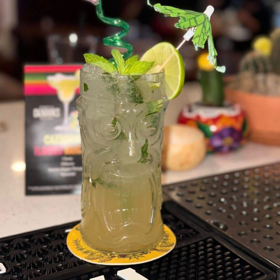 Mojito in a tiki glass with lime, mint, a green straw, and a small umbrella.