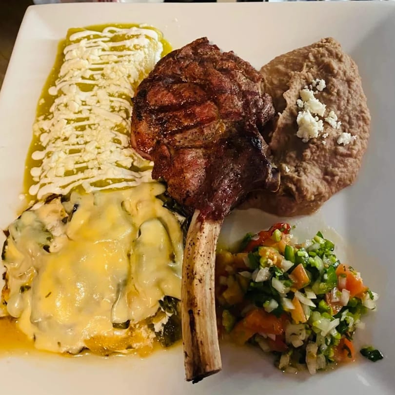 Grilled bone-in steak, green enchiladas, refried beans, chile relleno, and pico de gallo on a white plate.